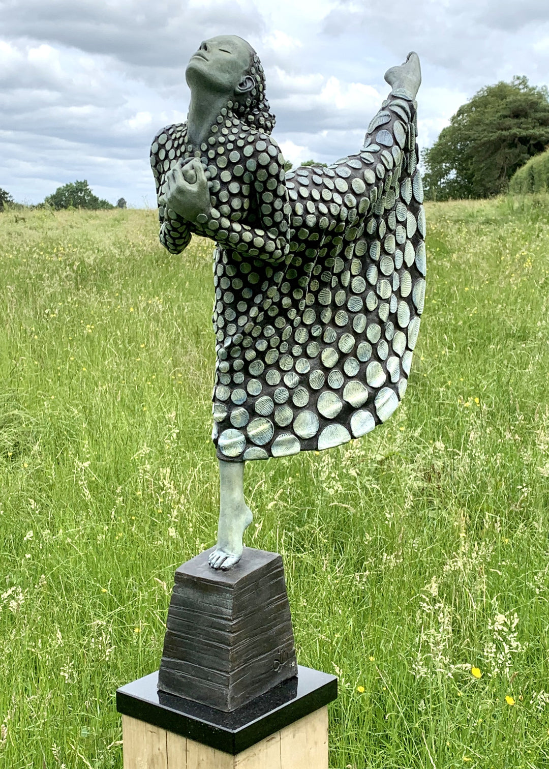 Bronze sculpture of a female figure poised on one leg atop a tiered plinth, her body arched back and arms drawn to her chest as she lifts her face toward the sky. Her dress and skin are patterned with circular, moon-like discs that flow outward into a crescent shape, photographed outdoors in a wide green landscape beneath a cloudy sky.