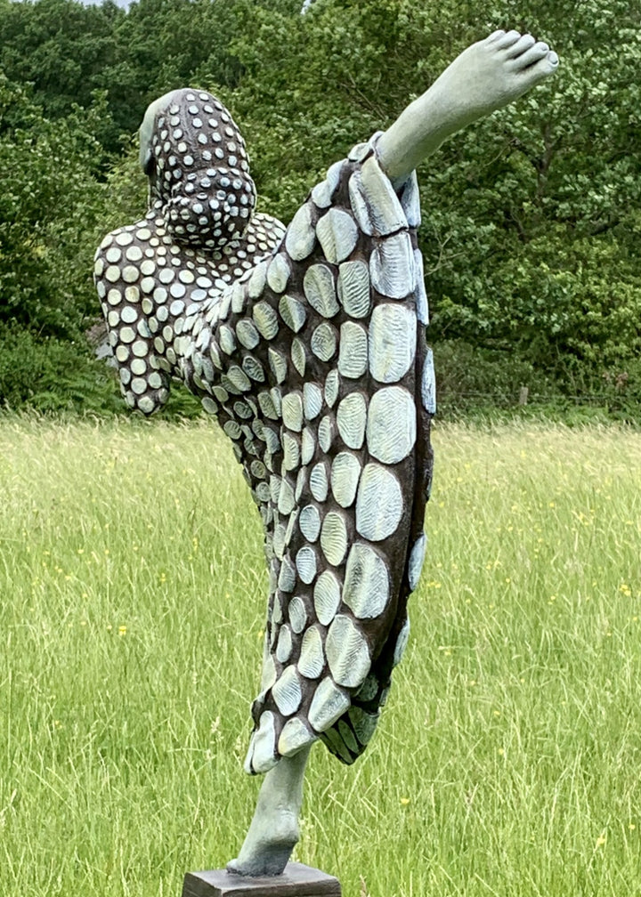 Bronze sculpture of a female figure poised on one leg atop a tiered plinth, her body arched back and arms drawn to her chest as she lifts her face toward the sky. Her dress and skin are patterned with circular, moon-like discs that flow outward into a crescent shape, photographed outdoors in a wide green landscape beneath a cloudy sky.