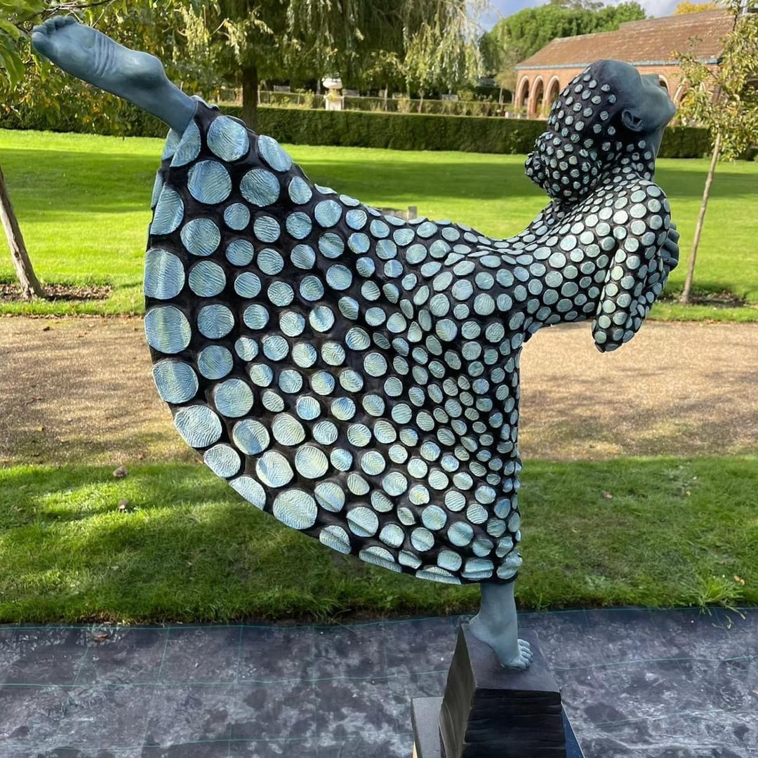 Bronze sculpture of a female figure poised on one leg atop a tiered plinth, her body arched back and arms drawn to her chest as she lifts her face toward the sky. Her dress and skin are patterned with circular, moon-like discs that flow outward into a crescent shape, photographed outdoors in a wide green landscape beneath a cloudy sky.