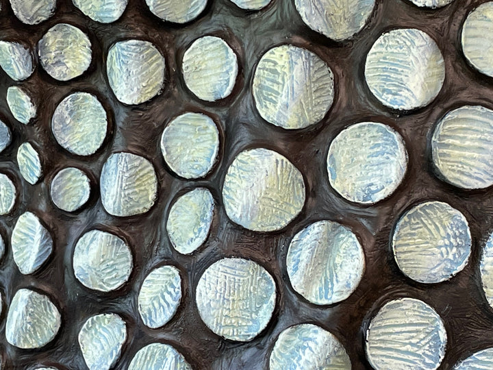 Bronze sculpture of a female figure poised on one leg atop a tiered plinth, her body arched back and arms drawn to her chest as she lifts her face toward the sky. Her dress and skin are patterned with circular, moon-like discs that flow outward into a crescent shape, photographed outdoors in a wide green landscape beneath a cloudy sky.