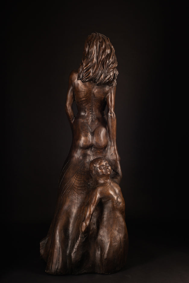 Tall figurative sculpture of a woman carved from richly grained wood, standing with her head lifted toward the sky. Her form emerges organically from the trunk, evoking growth and connection to the earth. The flowing grain patterns enhance her posture, while a small child figure is carved into the back of the sculpture, reaching upward and held by the woman's hand. A deeply symbolic piece that blends maternal strength, natural cycles, and generational continuity. The bronze preserves the texture of wood.