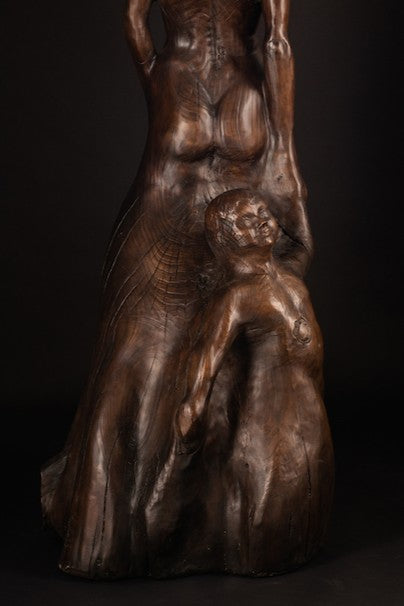 Tall figurative sculpture of a woman carved from richly grained wood, standing with her head lifted toward the sky. Her form emerges organically from the trunk, evoking growth and connection to the earth. The flowing grain patterns enhance her posture, while a small child figure is carved into the back of the sculpture, reaching upward and held by the woman's hand. A deeply symbolic piece that blends maternal strength, natural cycles, and generational continuity. The bronze preserves the texture of wood.