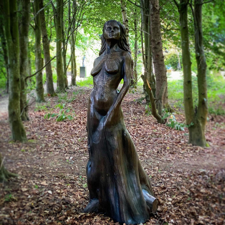 Tall figurative sculpture of a woman carved from richly grained wood, standing with her head lifted toward the sky. Her form emerges organically from the trunk, evoking growth and connection to the earth. The flowing grain patterns enhance her posture, while a small child figure is carved into the back of the sculpture, reaching upward and held by the woman's hand. A deeply symbolic piece that blends maternal strength, natural cycles, and generational continuity. The bronze preserves the texture of wood.