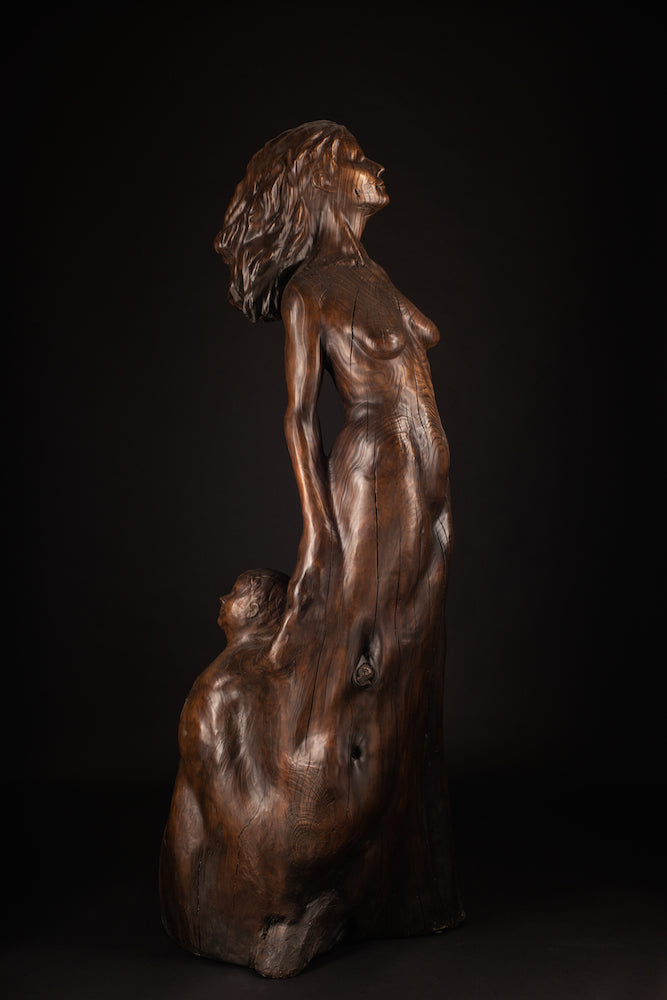 Tall figurative sculpture of a woman carved from richly grained wood, standing with her head lifted toward the sky. Her form emerges organically from the trunk, evoking growth and connection to the earth. The flowing grain patterns enhance her posture, while a small child figure is carved into the back of the sculpture, reaching upward and held by the woman's hand. A deeply symbolic piece that blends maternal strength, natural cycles, and generational continuity. The bronze preserves the texture of wood.