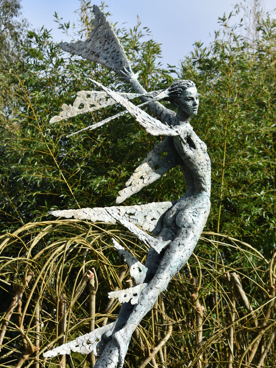 Monumental bronze sculpture of Theia, Greek goddess of light, portrayed in an elegant, elongated form with flowing limbs and wing-like extensions. Her textured surface and dynamic pose evoke movement and radiance, with light-catching shards projecting from her figure against open sky and landscape.