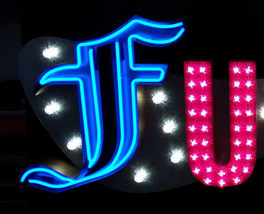 Chris Bracey, neon man, master of glow, gods own junkyard, Illuminated neon sign spelling out an expletive in bold, stylised letters — each letter lit in different colours and styles, including blue script, pink block lights, yellow cursive, and white marquee bulbs, mounted against a dark backdrop.