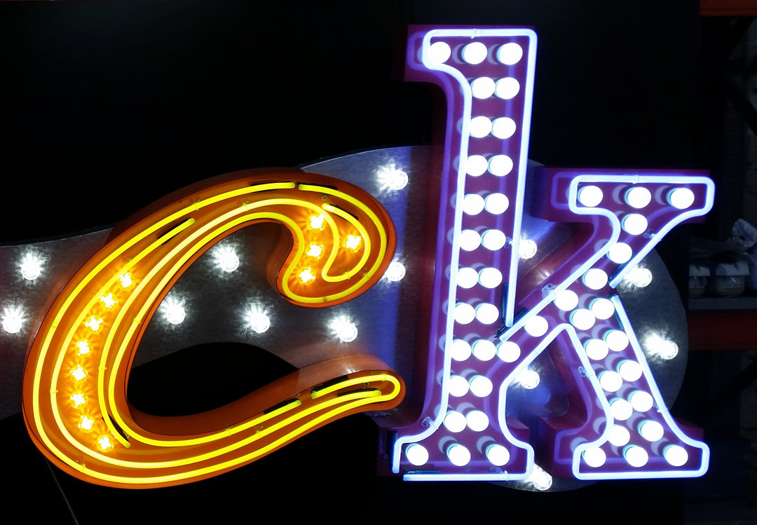 Chris Bracey, neon man, master of glow, gods own junkyard, Illuminated neon sign spelling out an expletive in bold, stylised letters — each letter lit in different colours and styles, including blue script, pink block lights, yellow cursive, and white marquee bulbs, mounted against a dark backdrop.