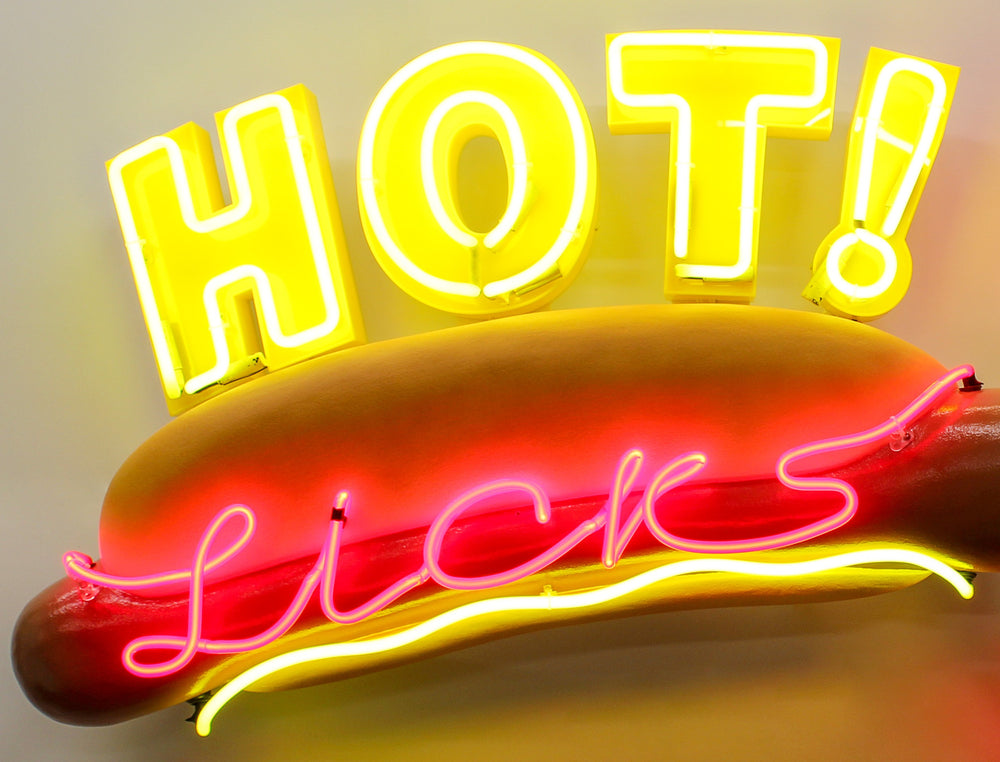 Playful neon wall art by Chris Bracey featuring a hot dog with the words “HOT Licks” in glowing yellow and pink neon, paired with a bold red neon mouth and lips design. Sculptural elements include a fabricated hot dog and 3D lips, combining humour and innuendo in classic Bracey style.