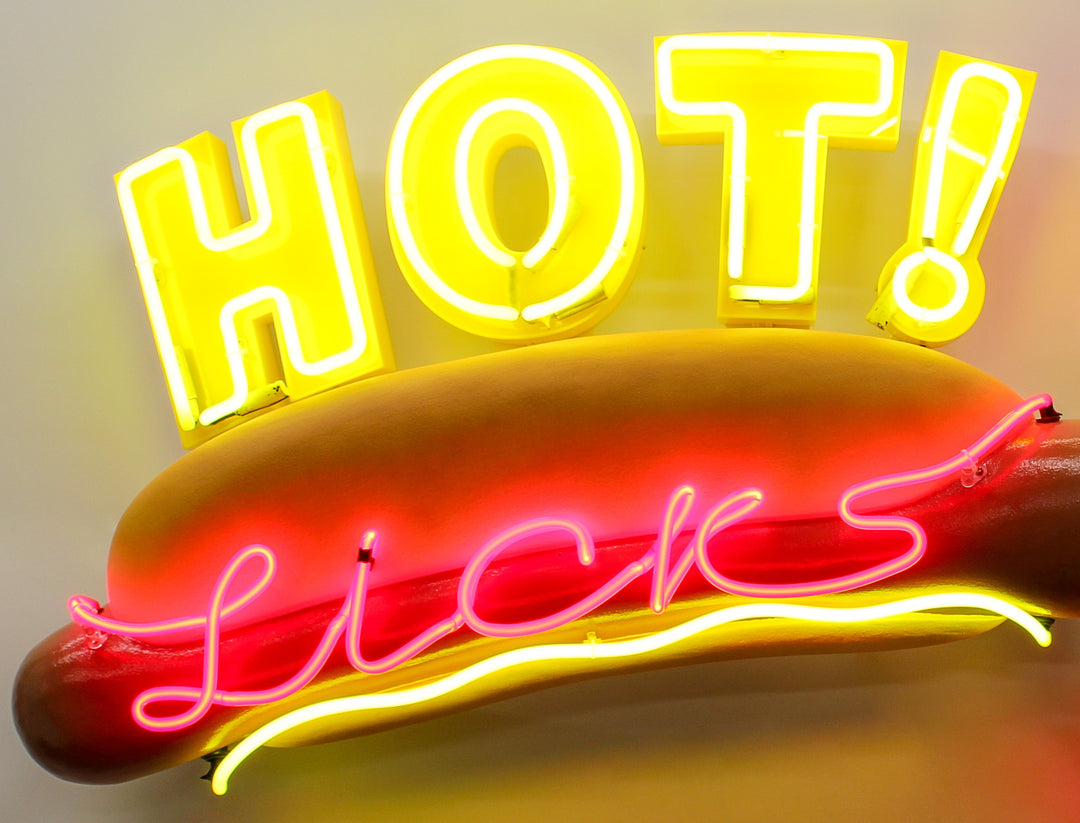 Playful neon wall art by Chris Bracey featuring a hot dog with the words “HOT Licks” in glowing yellow and pink neon, paired with a bold red neon mouth and lips design. Sculptural elements include a fabricated hot dog and 3D lips, combining humour and innuendo in classic Bracey style.