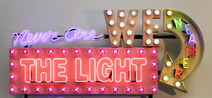 Neon wall artwork featuring the phrase “Never Are We Nearer The Light Than When The Darkness Is Deepest” in layered typography. Includes fabricated metal panels, red marquee lights, a retro arrow lit with cabochon LEDs, and vivid neon lettering in magenta, violet, red, blue, turquoise, orange, and white. A glowing, multicoloured composition by Chris Bracey reflecting spiritual insight and emotional depth.