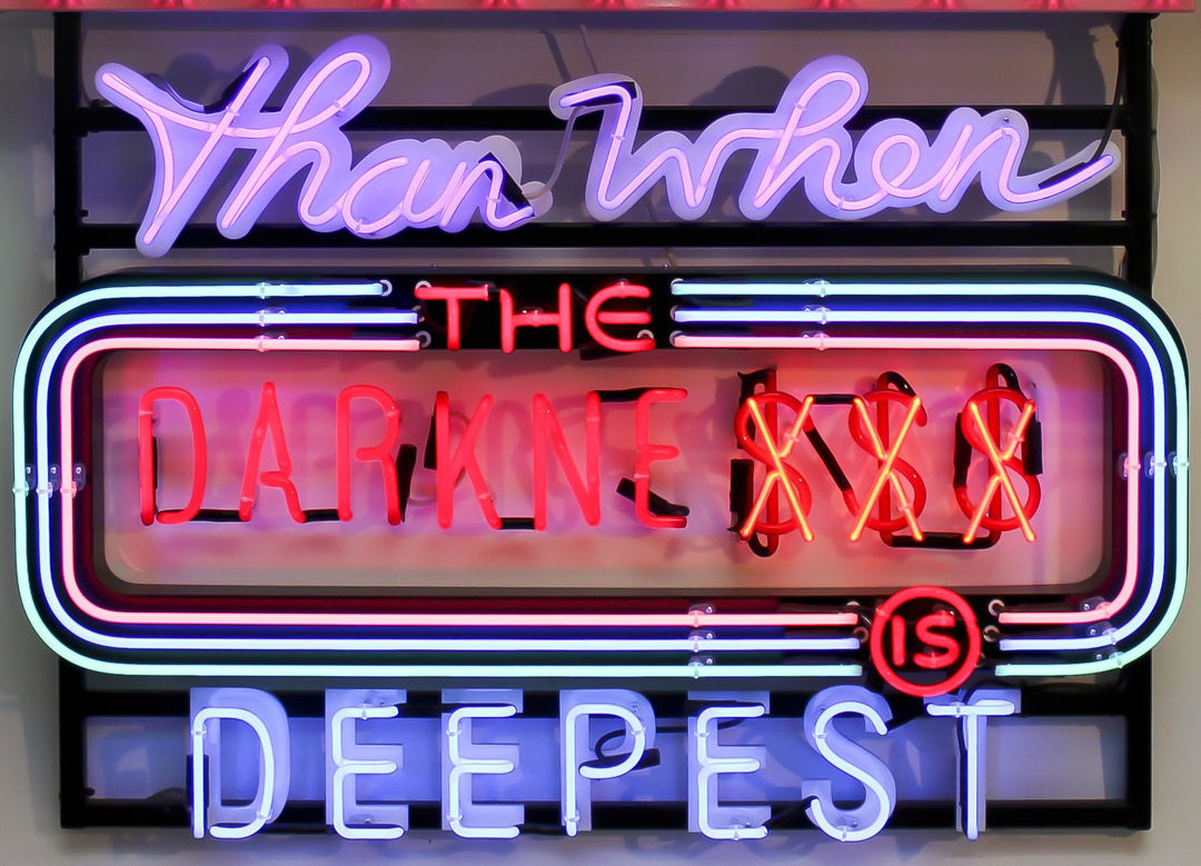 Neon wall artwork featuring the phrase “Never Are We Nearer The Light Than When The Darkness Is Deepest” in layered typography. Includes fabricated metal panels, red marquee lights, a retro arrow lit with cabochon LEDs, and vivid neon lettering in magenta, violet, red, blue, turquoise, orange, and white. A glowing, multicoloured composition by Chris Bracey reflecting spiritual insight and emotional depth.