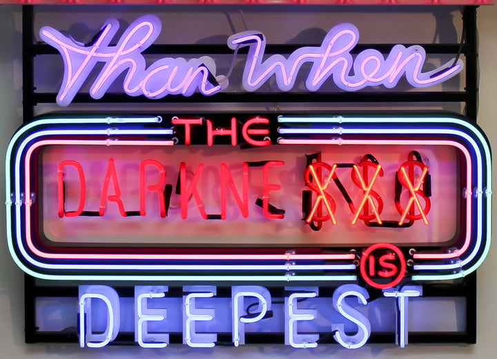 Neon wall artwork featuring the phrase “Never Are We Nearer The Light Than When The Darkness Is Deepest” in layered typography. Includes fabricated metal panels, red marquee lights, a retro arrow lit with cabochon LEDs, and vivid neon lettering in magenta, violet, red, blue, turquoise, orange, and white. A glowing, multicoloured composition by Chris Bracey reflecting spiritual insight and emotional depth.