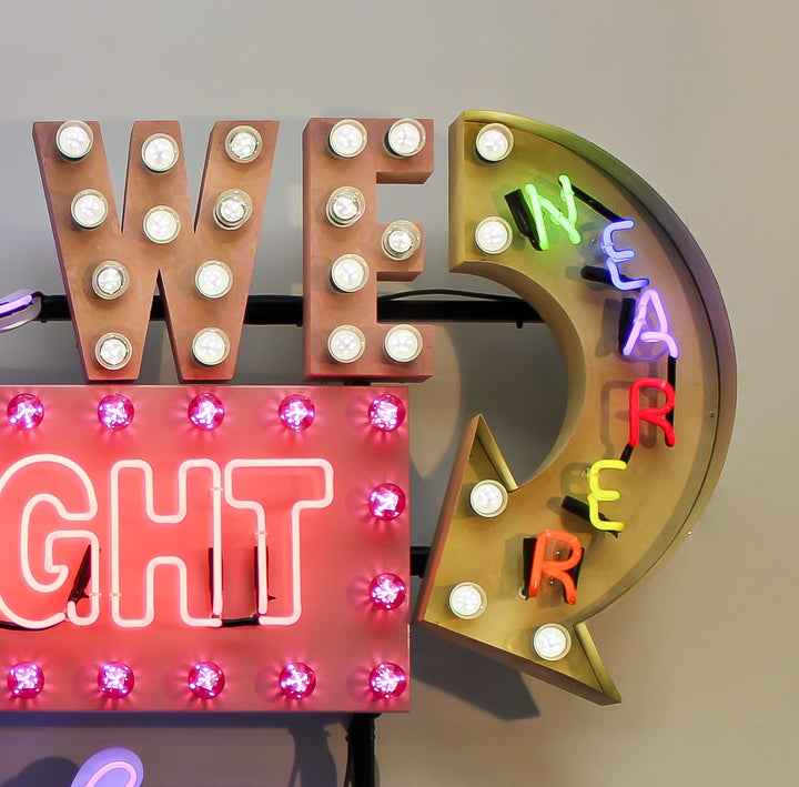 Neon wall artwork featuring the phrase “Never Are We Nearer The Light Than When The Darkness Is Deepest” in layered typography. Includes fabricated metal panels, red marquee lights, a retro arrow lit with cabochon LEDs, and vivid neon lettering in magenta, violet, red, blue, turquoise, orange, and white. A glowing, multicoloured composition by Chris Bracey reflecting spiritual insight and emotional depth.