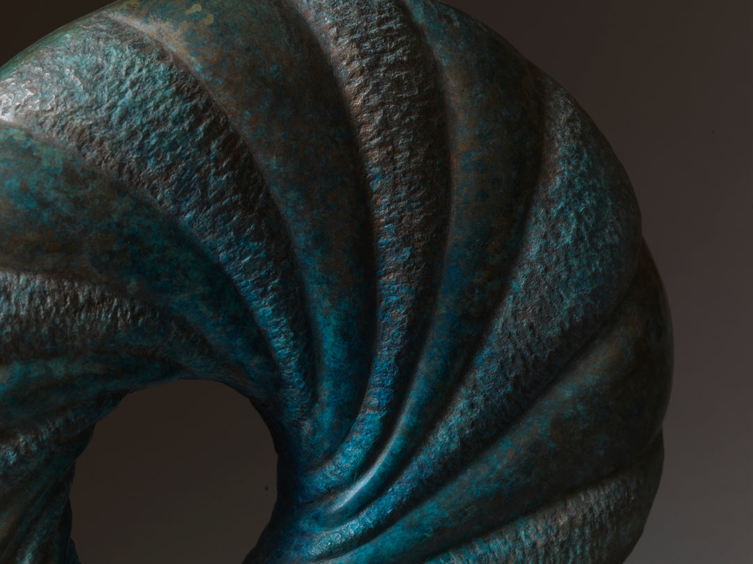‘Praa’ by Nicolas Moreton MRSS, a limited-edition contemporary abstract bronze sculpture featuring a circular form with a central opening and deeply carved spiralling ridges. Alternating smooth and textured surfaces are finished in turquoise, teal, blue-green and dark bronze patina, creating a twisting pattern around the open centre. The sculpture is displayed on a rectangular pale cream stone base against a dark grey and brown studio background.