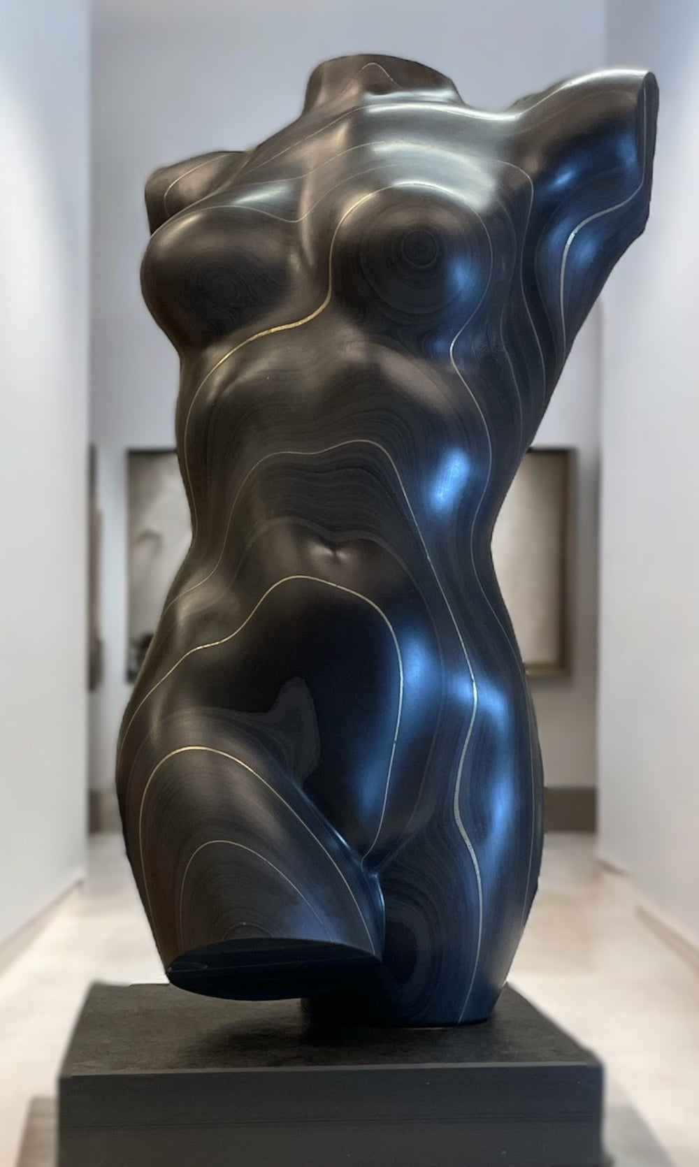 Sculpted female torso in smooth black slate with natural striations and gold-toned contour lines, mounted on a dark plinth and displayed in a modern interior setting.