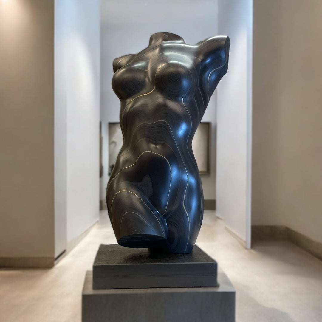 Sculpted female torso in smooth black slate with natural striations and gold-toned contour lines, mounted on a dark plinth and displayed in a modern interior setting.