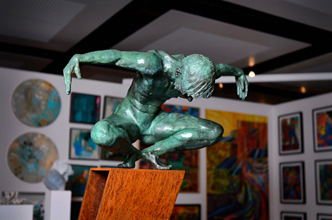 Large bronze sculpture of a crouching male figure, arms extended and head bowed, balanced on the edge of an angled rusted steel plinth. The figure’s green patinated surface contrasts with the warm, textured tones of the steel base. Muscular detail and tension in the pose evoke focus, strength, and introspection. Displayed both indoors against gallery walls and outdoors in a garden setting, the work captures light and shadow across the form, emphasising anatomical realism and expressive movement.