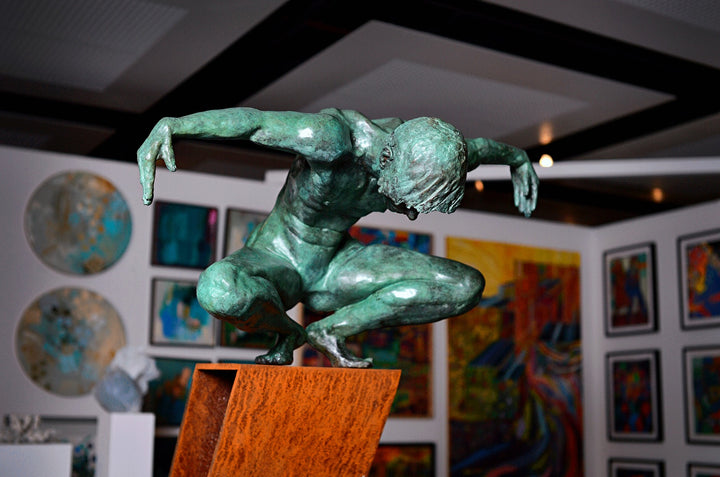 Large bronze sculpture of a crouching male figure, arms extended and head bowed, balanced on the edge of an angled rusted steel plinth. The figure’s green patinated surface contrasts with the warm, textured tones of the steel base. Muscular detail and tension in the pose evoke focus, strength, and introspection. Displayed both indoors against gallery walls and outdoors in a garden setting, the work captures light and shadow across the form, emphasising anatomical realism and expressive movement.