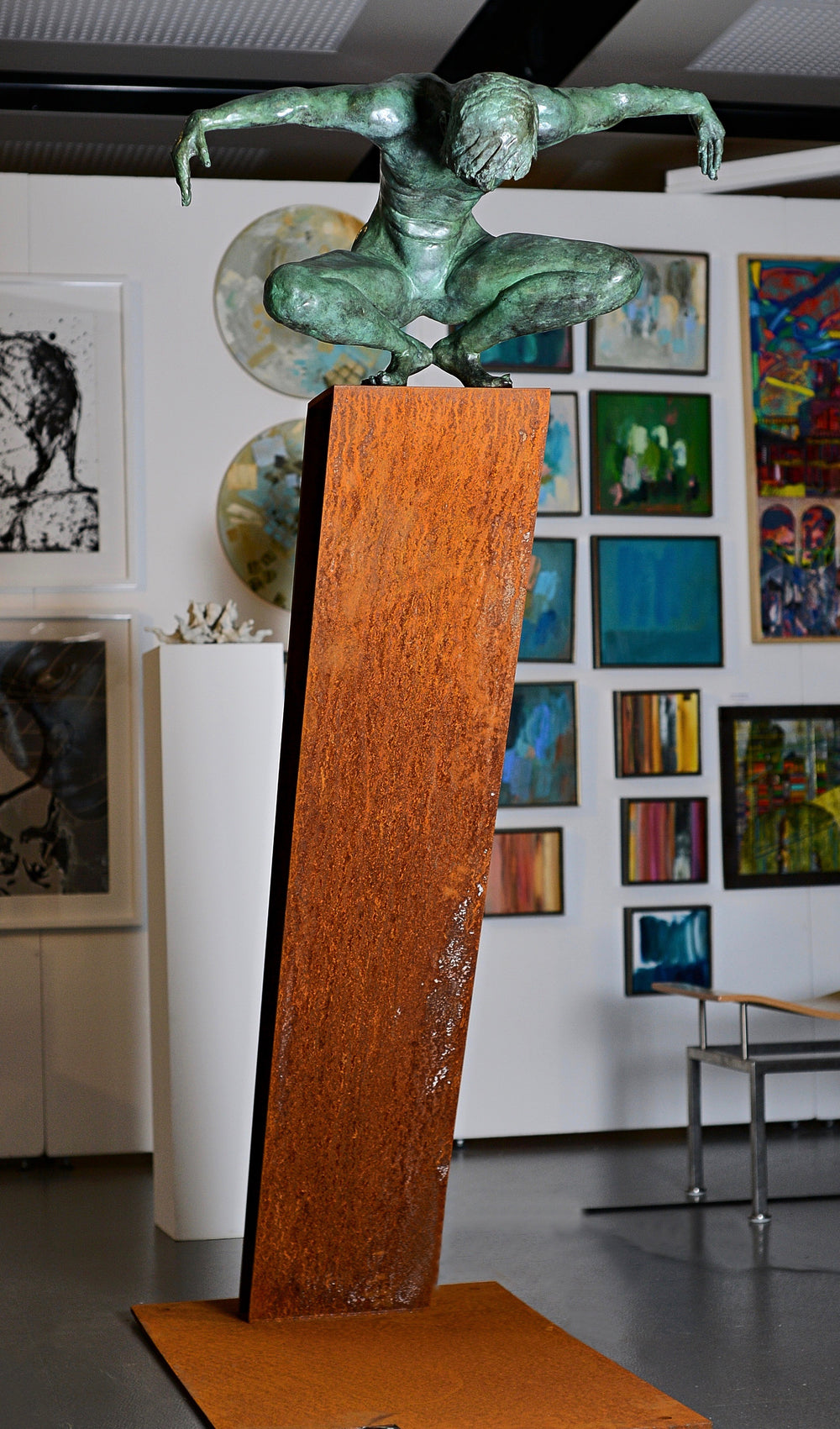 Large bronze sculpture of a crouching male figure, arms extended and head bowed, balanced on the edge of an angled rusted steel plinth. The figure’s green patinated surface contrasts with the warm, textured tones of the steel base. Muscular detail and tension in the pose evoke focus, strength, and introspection. Displayed both indoors against gallery walls and outdoors in a garden setting, the work captures light and shadow across the form, emphasising anatomical realism and expressive movement.