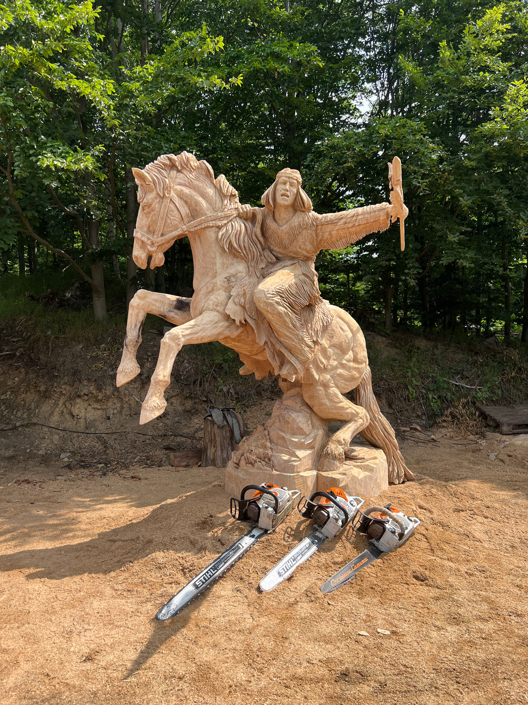 Imposing chainsaw-carved wooden sculpture of a Native American warrior riding a rearing horse, arm outstretched holding a ceremonial object, set against a woodland backdrop with sawdust and chainsaws visible in the foreground.