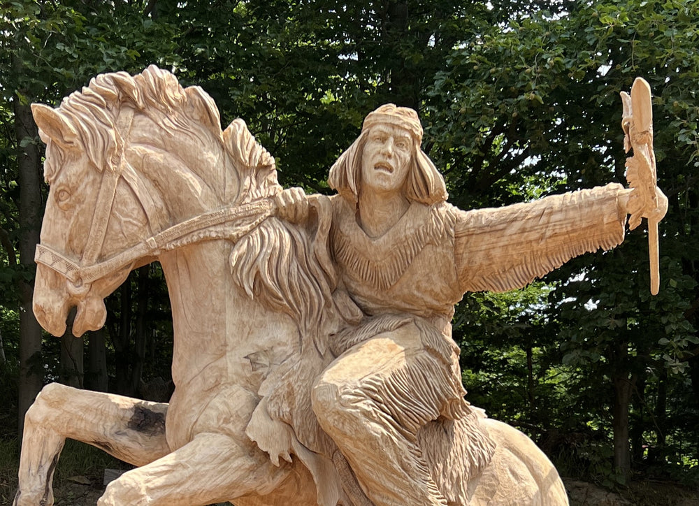 Imposing chainsaw-carved wooden sculpture of a Native American warrior riding a rearing horse, arm outstretched holding a ceremonial object, set against a woodland backdrop with sawdust and chainsaws visible in the foreground.