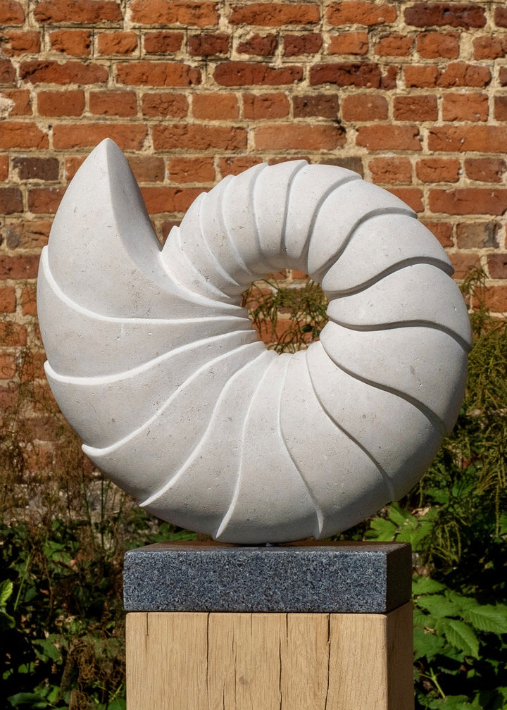 Nautilus by Nicola Axe — elegant spiral Portland stone sculpture echoing a seashell’s natural form, smooth pale surface with flowing ridges, mounted on a granite and oak plinth with a discreet metal plate.