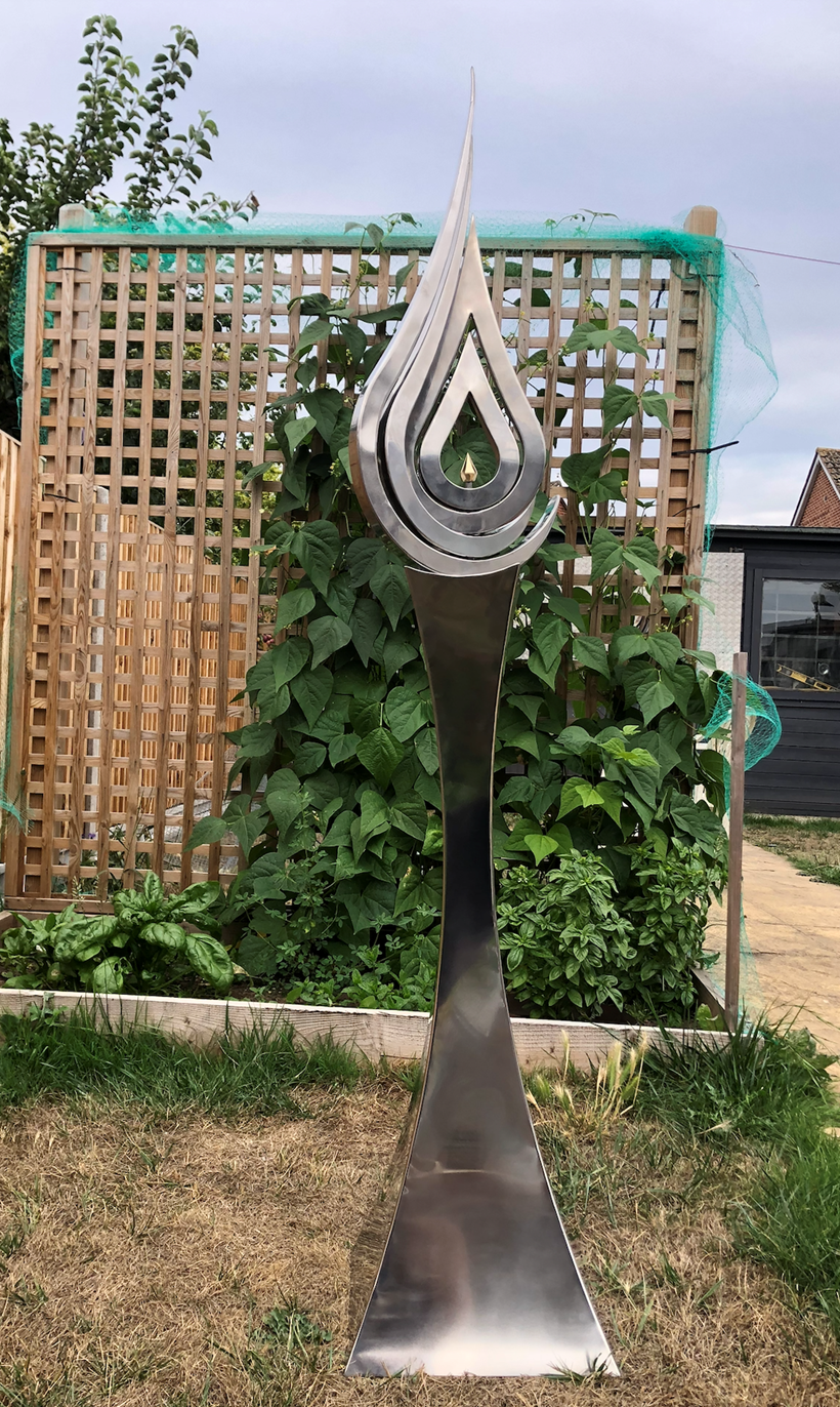 An elegant stainless steel kinetic sculpture by Martin Jarvis, designed in the form of a tall flame-like spire. The sculpture’s upper structure features fluid, ribbon-like curves that twist upward and move gently with the wind. Polished to a mirror finish, the steel reflects light and surroundings, giving it a dynamic presence. The base is solid and tapering, with soft surface patterning that adds depth and texture. Installed outdoors in natural and garden settings, the piece feels both modern 