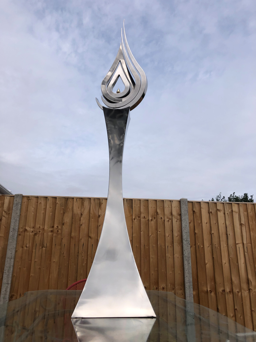 An elegant stainless steel kinetic sculpture by Martin Jarvis, designed in the form of a tall flame-like spire. The sculpture’s upper structure features fluid, ribbon-like curves that twist upward and move gently with the wind. Polished to a mirror finish, the steel reflects light and surroundings, giving it a dynamic presence. The base is solid and tapering, with soft surface patterning that adds depth and texture. Installed outdoors in natural and garden settings, the piece feels both modern 