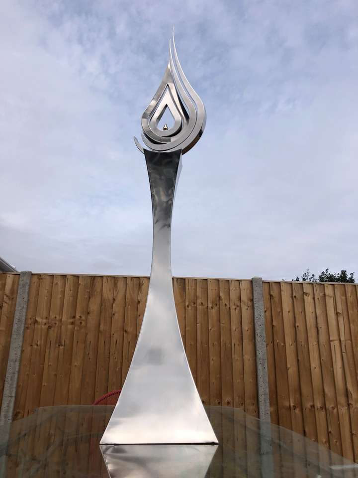 An elegant stainless steel kinetic sculpture by Martin Jarvis, designed in the form of a tall flame-like spire. The sculpture’s upper structure features fluid, ribbon-like curves that twist upward and move gently with the wind. Polished to a mirror finish, the steel reflects light and surroundings, giving it a dynamic presence. The base is solid and tapering, with soft surface patterning that adds depth and texture. Installed outdoors in natural and garden settings, the piece feels both modern 