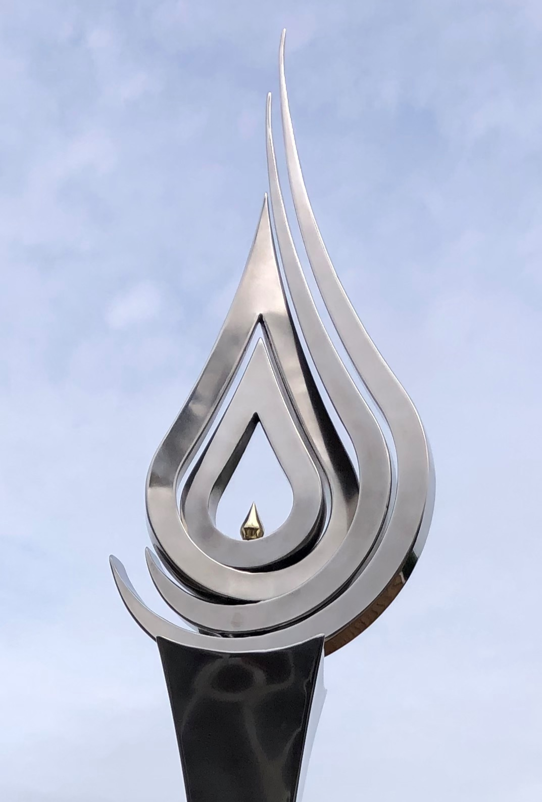 An elegant stainless steel kinetic sculpture by Martin Jarvis, designed in the form of a tall flame-like spire. The sculpture’s upper structure features fluid, ribbon-like curves that twist upward and move gently with the wind. Polished to a mirror finish, the steel reflects light and surroundings, giving it a dynamic presence. The base is solid and tapering, with soft surface patterning that adds depth and texture. Installed outdoors in natural and garden settings, the piece feels both modern 