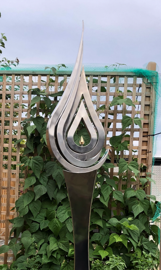 An elegant stainless steel kinetic sculpture by Martin Jarvis, designed in the form of a tall flame-like spire. The sculpture’s upper structure features fluid, ribbon-like curves that twist upward and move gently with the wind. Polished to a mirror finish, the steel reflects light and surroundings, giving it a dynamic presence. The base is solid and tapering, with soft surface patterning that adds depth and texture. Installed outdoors in natural and garden settings, the piece feels both modern 