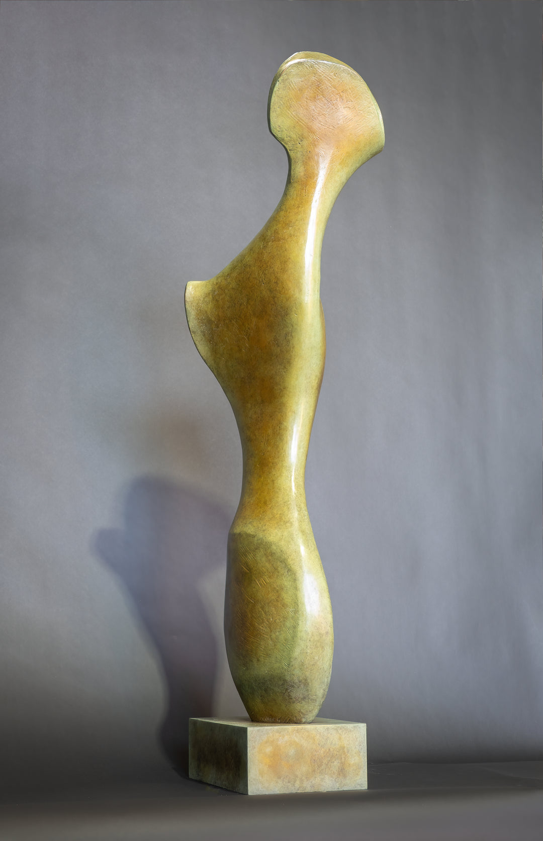 Bone Form IV — abstract bronze sculpture by Nicola Godden inspired by the natural curves and hollows of animal bone. The piece has a tall, sinuous form with a hollowed centre, organic surface texture and warm bronze patina shifting from earthy brown to soft green and gold highlights. Displayed outdoors on a one-metre timber plinth against a bright blue sky and winter landscape.