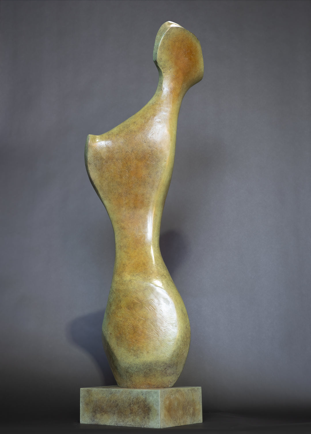 Bone Form IV — abstract bronze sculpture by Nicola Godden inspired by the natural curves and hollows of animal bone. The piece has a tall, sinuous form with a hollowed centre, organic surface texture and warm bronze patina shifting from earthy brown to soft green and gold highlights. Displayed outdoors on a one-metre timber plinth against a bright blue sky and winter landscape.