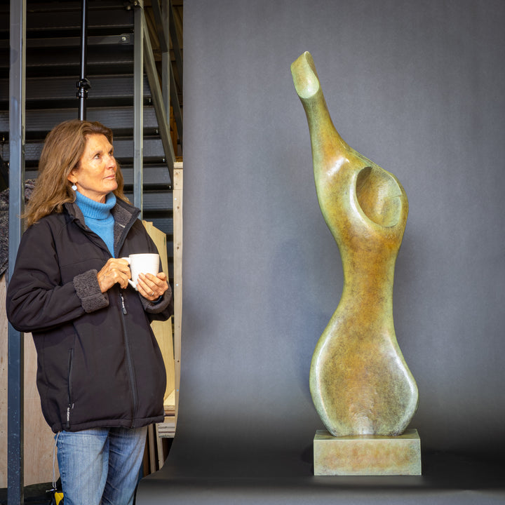 Bone Form IV — abstract bronze sculpture by Nicola Godden inspired by the natural curves and hollows of animal bone. The piece has a tall, sinuous form with a hollowed centre, organic surface texture and warm bronze patina shifting from earthy brown to soft green and gold highlights. Displayed outdoors on a one-metre timber plinth against a bright blue sky and winter landscape.