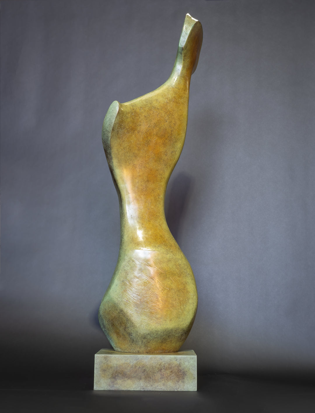 Bone Form IV — abstract bronze sculpture by Nicola Godden inspired by the natural curves and hollows of animal bone. The piece has a tall, sinuous form with a hollowed centre, organic surface texture and warm bronze patina shifting from earthy brown to soft green and gold highlights. Displayed outdoors on a one-metre timber plinth against a bright blue sky and winter landscape.