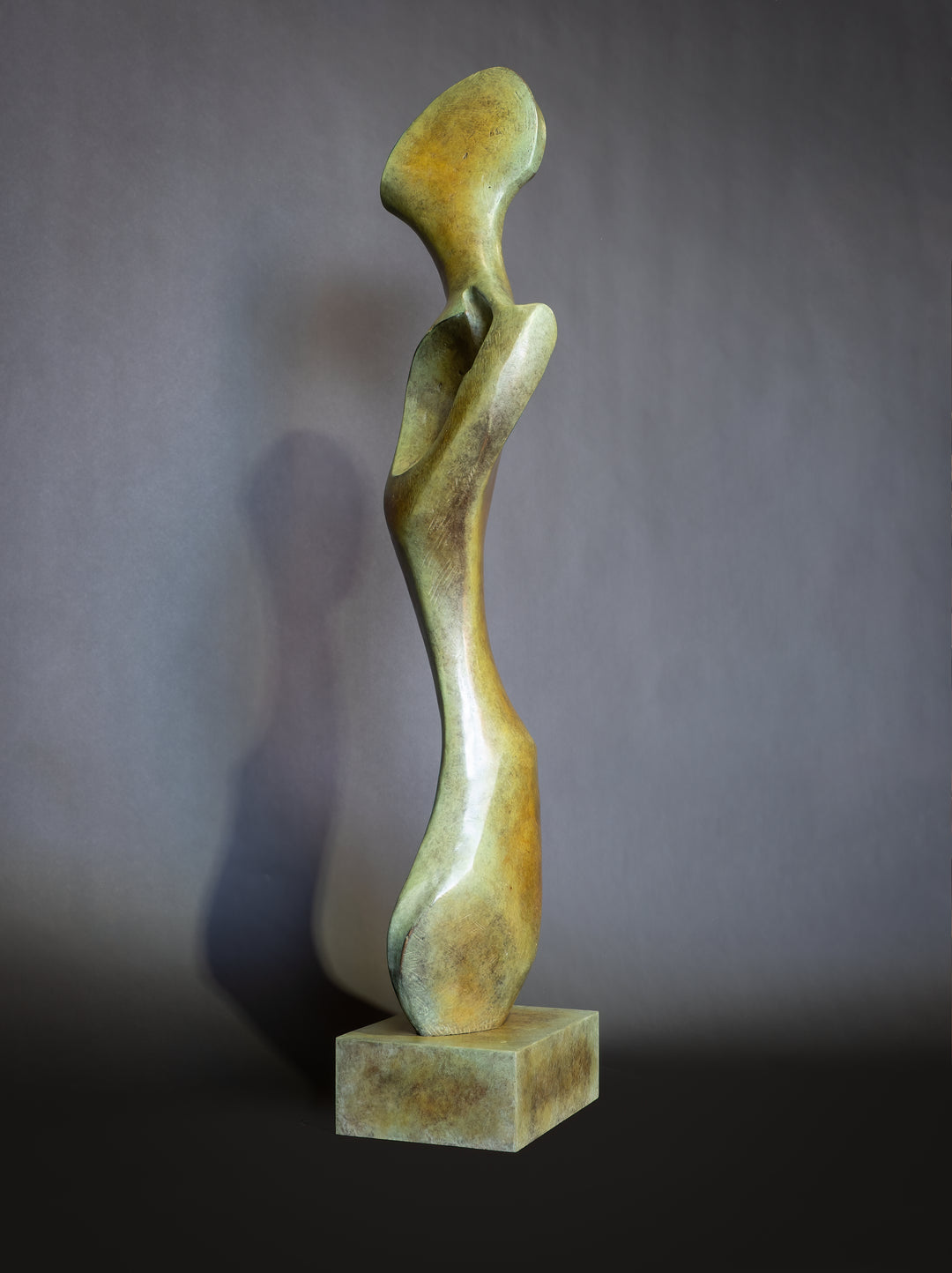 Bone Form IV — abstract bronze sculpture by Nicola Godden inspired by the natural curves and hollows of animal bone. The piece has a tall, sinuous form with a hollowed centre, organic surface texture and warm bronze patina shifting from earthy brown to soft green and gold highlights. Displayed outdoors on a one-metre timber plinth against a bright blue sky and winter landscape.