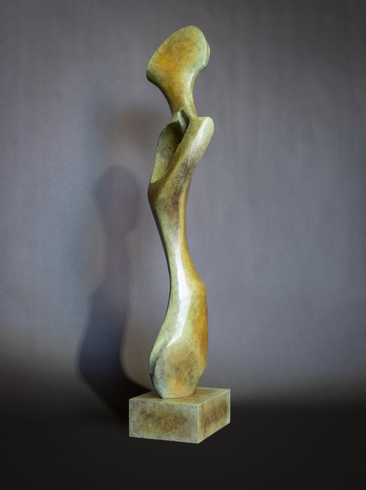 Bone Form IV — abstract bronze sculpture by Nicola Godden inspired by the natural curves and hollows of animal bone. The piece has a tall, sinuous form with a hollowed centre, organic surface texture and warm bronze patina shifting from earthy brown to soft green and gold highlights. Displayed outdoors on a one-metre timber plinth against a bright blue sky and winter landscape.