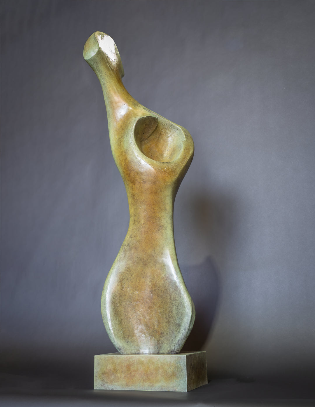 Bone Form IV — abstract bronze sculpture by Nicola Godden inspired by the natural curves and hollows of animal bone. The piece has a tall, sinuous form with a hollowed centre, organic surface texture and warm bronze patina shifting from earthy brown to soft green and gold highlights. Displayed outdoors on a one-metre timber plinth against a bright blue sky and winter landscape.