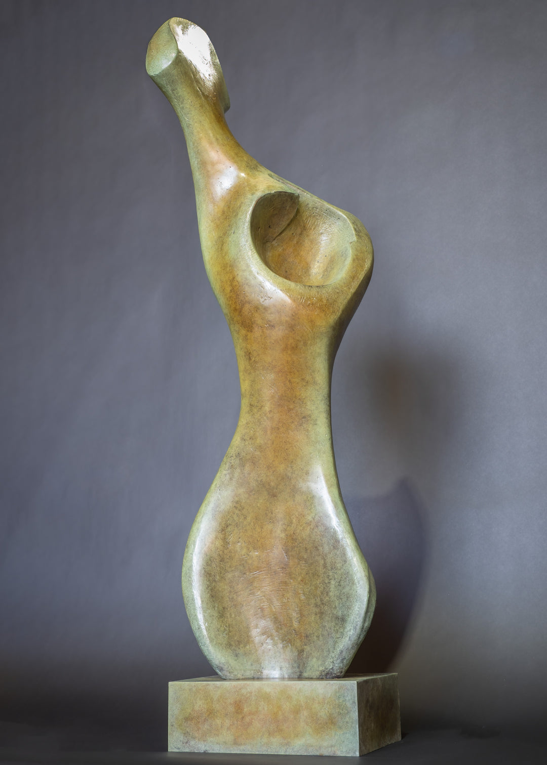 Bone Form IV — abstract bronze sculpture by Nicola Godden inspired by the natural curves and hollows of animal bone. The piece has a tall, sinuous form with a hollowed centre, organic surface texture and warm bronze patina shifting from earthy brown to soft green and gold highlights. Displayed outdoors on a one-metre timber plinth against a bright blue sky and winter landscape.