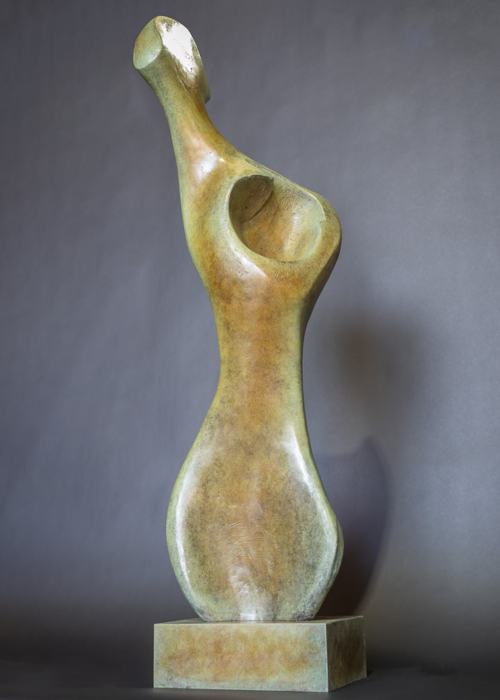 Bone Form IV — abstract bronze sculpture by Nicola Godden inspired by the natural curves and hollows of animal bone. The piece has a tall, sinuous form with a hollowed centre, organic surface texture and warm bronze patina shifting from earthy brown to soft green and gold highlights. Displayed outdoors on a one-metre timber plinth against a bright blue sky and winter landscape.