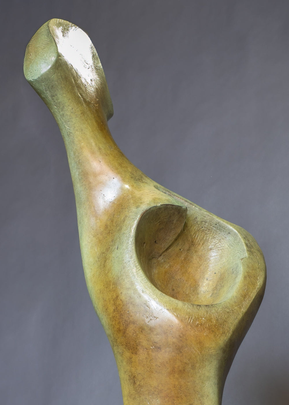 Bone Form IV — abstract bronze sculpture by Nicola Godden inspired by the natural curves and hollows of animal bone. The piece has a tall, sinuous form with a hollowed centre, organic surface texture and warm bronze patina shifting from earthy brown to soft green and gold highlights. Displayed outdoors on a one-metre timber plinth against a bright blue sky and winter landscape.