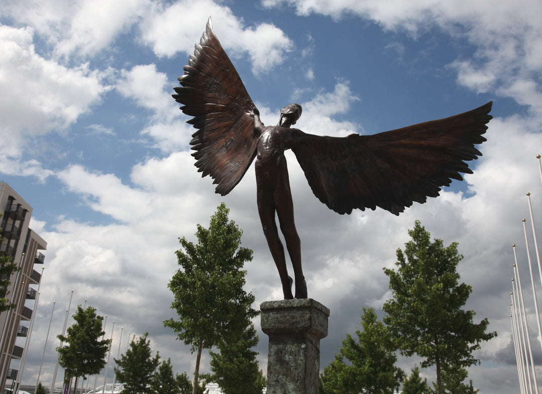 Icarus I — monumental bronze sculpture by Nicola Godden, depicting a stylised male figure with immense outstretched wings, leaning forward as if preparing for flight. Rich bronze patina with warm coppery browns fading into lighter golden tips across the feathered wings. Originally featured at the London 2012 Olympic Village and sold at Sotheby’s. Set outdoors against a clear blue sky and classical architectural backdrop.
