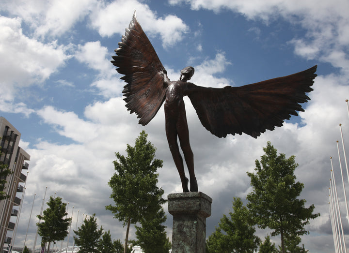 Icarus I — monumental bronze sculpture by Nicola Godden, depicting a stylised male figure with immense outstretched wings, leaning forward as if preparing for flight. Rich bronze patina with warm coppery browns fading into lighter golden tips across the feathered wings. Originally featured at the London 2012 Olympic Village and sold at Sotheby’s. Set outdoors against a clear blue sky and classical architectural backdrop.