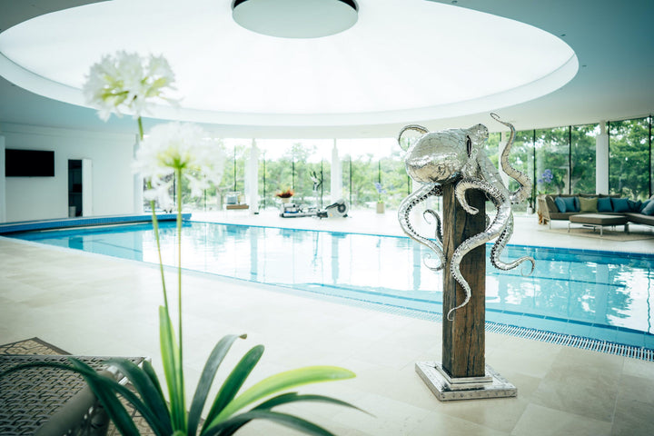 ‘Octopus’ by Michael Turner, a unique, one-of-one stainless steel marine wildlife sculpture depicting a large octopus perched on a tall, weathered hardwood post. Handcrafted from intricately shaped and welded stainless steel, the sculpture features a polished silver finish, a rounded head, detailed eyes and eight flowing tentacles with individually formed suction cups, curling around the wooden support. A small metallic crab climbs the post below, adding a playful detail. Mounted on a square stainless steel