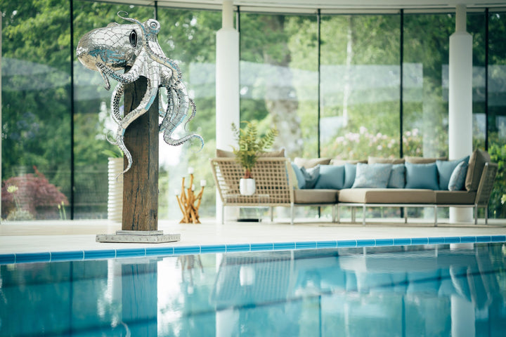 ‘Octopus’ by Michael Turner, a unique, one-of-one stainless steel marine wildlife sculpture depicting a large octopus perched on a tall, weathered hardwood post. Handcrafted from intricately shaped and welded stainless steel, the sculpture features a polished silver finish, a rounded head, detailed eyes and eight flowing tentacles with individually formed suction cups, curling around the wooden support. A small metallic crab climbs the post below, adding a playful detail. Mounted on a square stainless steel