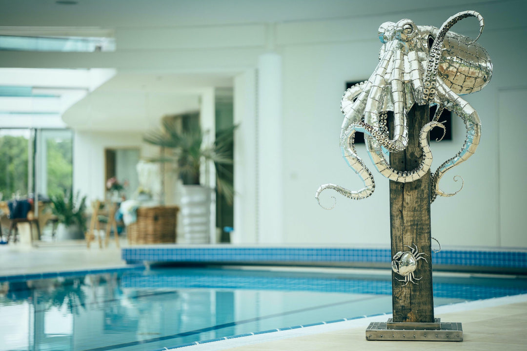 ‘Octopus’ by Michael Turner, a unique, one-of-one stainless steel marine wildlife sculpture depicting a large octopus perched on a tall, weathered hardwood post. Handcrafted from intricately shaped and welded stainless steel, the sculpture features a polished silver finish, a rounded head, detailed eyes and eight flowing tentacles with individually formed suction cups, curling around the wooden support. A small metallic crab climbs the post below, adding a playful detail. Mounted on a square stainless steel
