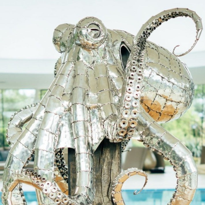 ‘Octopus’ by Michael Turner, a unique, one-of-one stainless steel marine wildlife sculpture depicting a large octopus perched on a tall, weathered hardwood post. Handcrafted from intricately shaped and welded stainless steel, the sculpture features a polished silver finish, a rounded head, detailed eyes and eight flowing tentacles with individually formed suction cups, curling around the wooden support. A small metallic crab climbs the post below, adding a playful detail. Mounted on a square stainless steel