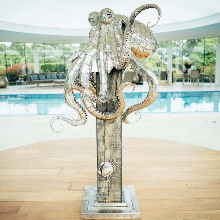 ‘Octopus’ by Michael Turner, a unique, one-of-one stainless steel marine wildlife sculpture depicting a large octopus perched on a tall, weathered hardwood post. Handcrafted from intricately shaped and welded stainless steel, the sculpture features a polished silver finish, a rounded head, detailed eyes and eight flowing tentacles with individually formed suction cups, curling around the wooden support. A small metallic crab climbs the post below, adding a playful detail. Mounted on a square stainless steel