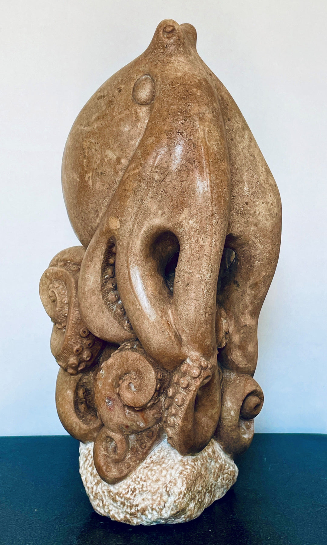 Octopus sculpture by Nicola Axe — intricately carved from warm pink serpentine, with coiled curling tentacles and smooth rounded head, perched on a pale textured stone base, blending marine life detail with organic elegance.