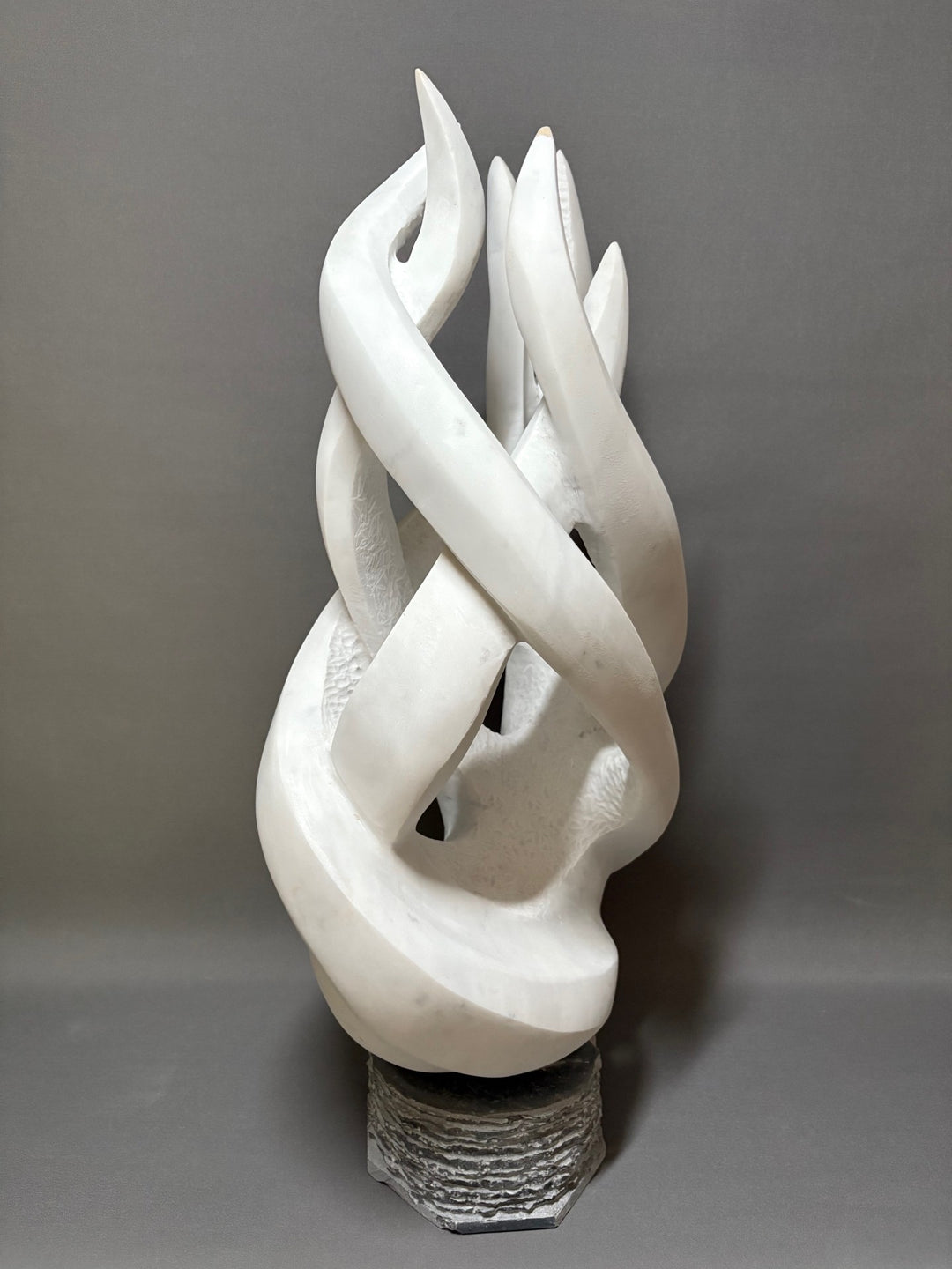 Once More by Brendan Cullen, a dynamic hand-carved sculpture in Carrara marble, formed of interlacing, flame-like ribbons that twist upward in fluid motion. The smooth white marble is contrasted with textured inner surfaces, giving depth and vitality to the piece. Mounted on a dark stone base, it captures a sense of energy, renewal, and continuous flow.